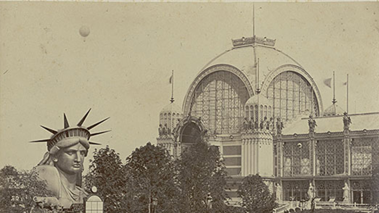 Photographie sépia qui montre la tête de statue de la Liberté, qui émerge des arbres, et le dôme en verrière de la Grande Galerie, en arrière-plan..