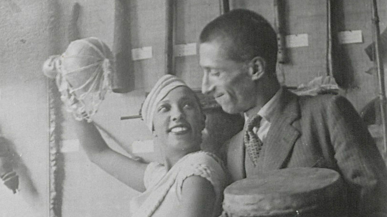 Photo en noir et blanc montrant Joséphine Baker, habillée de blanc, qui pose de manière espiègle avec un homme en costume, devant la vitrine d'une expo. Ils ont l'air complices et de s'amuser. Elle tien en main un instrument de musique africain et le monsieur un tambour.