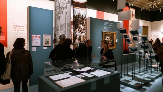 Dans une grande salle, installations de documents, tableaux et objets avec du public que l'on aperçoit dans la pénombre. 
