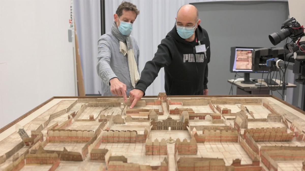 Deux hommes avec des masques hygiéniques, le photographe et l’ingénieur, sont debout. Ils se tiennent devant un grand plan en relief ancien. ils montrent du doigt un bâtiment de la maquette. À côté d'eux, se trouve l'appareil photo numérique. 