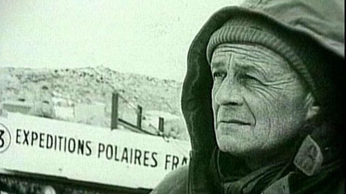 Photo en noir et blanc montrant Paul- Emile Victor de profil, habillé en tenue polaire, avec en fond un fronton de bâtiment où il est indiqué "Expéditions polaires françaises". 