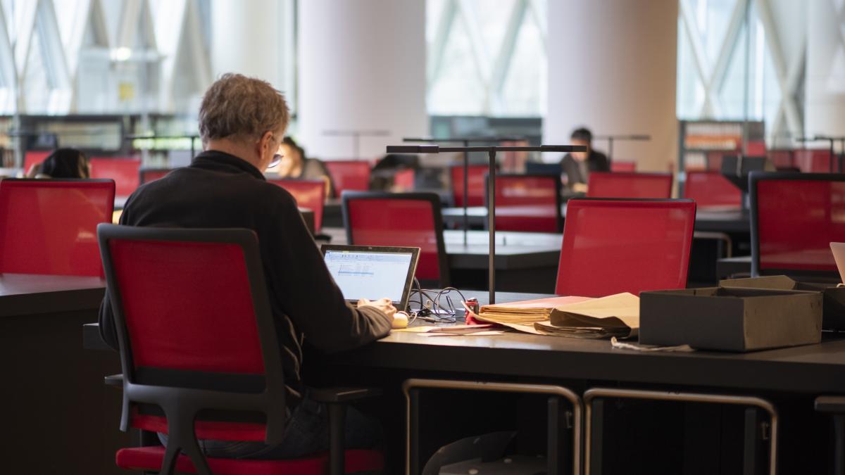 Salle de lecture du site de Pierrefitte-sur-Seine © Carole Bauer / Archives nationales de France Vue de dos d'un lecteur devant son ordi portable en train de consulter des documents. Au second plan, les alignements des tables et chaises rouges de la vaste salle.