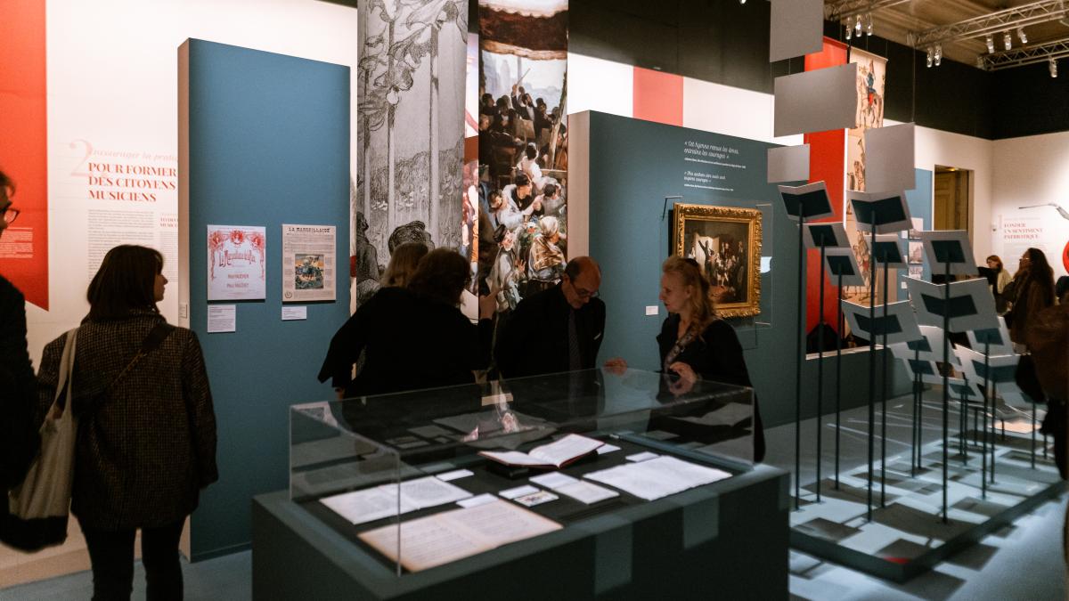 Dans une grande salle, installations de documents, tableaux et objets avec du public que l'on aperçoit dans la pénombre. 