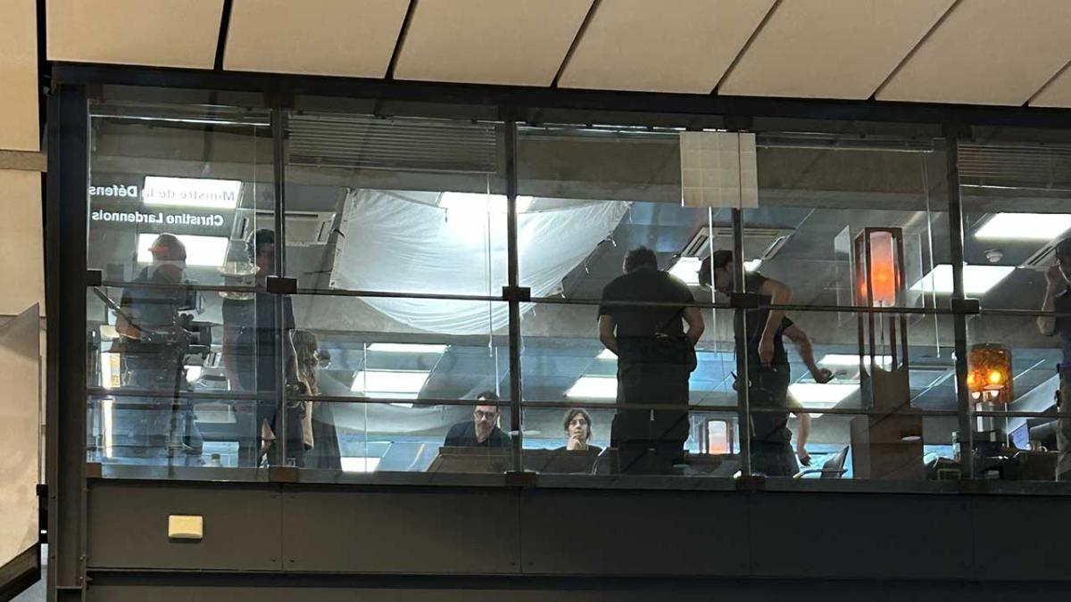 Bureaux avec de grandes cloisons de verre. On aperçoit des silhouettes de l'équipe de tournage à travers.