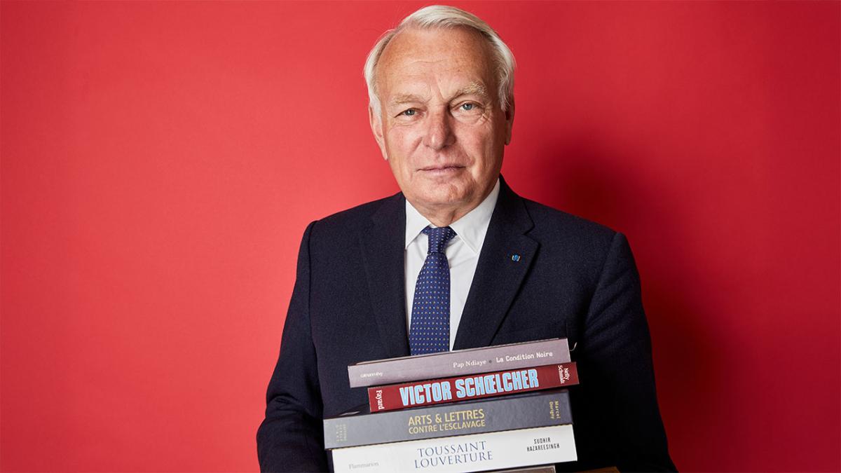Portrait en buste de Jean-Marc Ayrault. Il est de face, devant un fond rouge et il tient une pile de livres sur l'histoire de l'esclavage. 