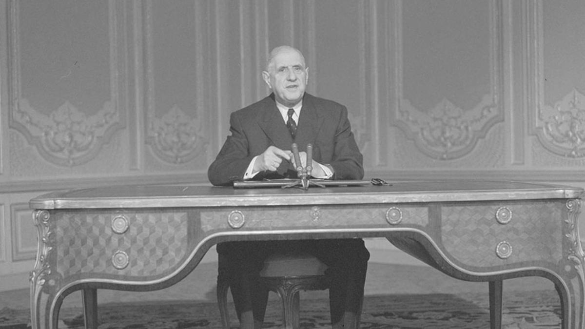 Photo en noir et blanc montrant de Gaulle, en costume cravate, assis derrière un grand bureau de mobilier ancien, avec 2 micros devant lui. 