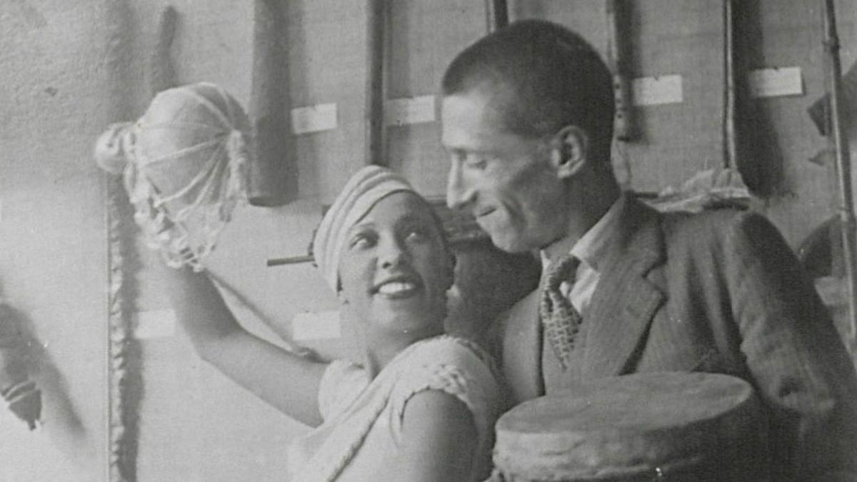 Photo en noir et blanc montrant Joséphine Baker, habillée de blanc, qui pose de manière espiègle avec un homme en costume, devant la vitrine d'une expo. Ils ont l'air complices et de s'amuser. Elle tien en main un instrument de musique africain et le monsieur un tambour.