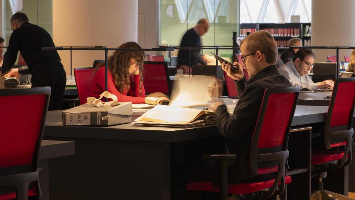 Salle de lecture du site de Pierrefitte-sur-Seine Assis dans des fauteuils rouges, des adultes consultent des documents d'archives.