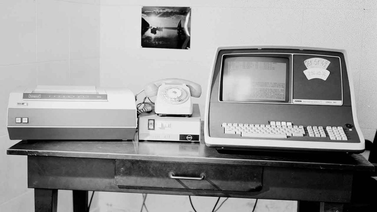 « Matériel ancien : Leonor. Ordinateur » de 1979 Sur une table en bois devant un mur décoré d'une photo, un très gros ordinateur de 1979, doté d'un petit écran et d'un gros clavier. À gauche, une sorte d'imprimante portative et, au milieu, un téléphone blanc à combiné et cadran. La photo est en noir et blanc.