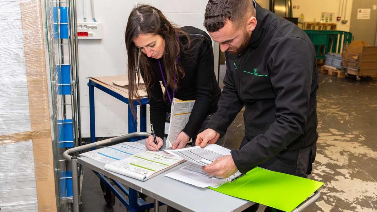 Une femme et un homme sont penchés sur des documents dans un entrepôt, pour vérifier une liste.  