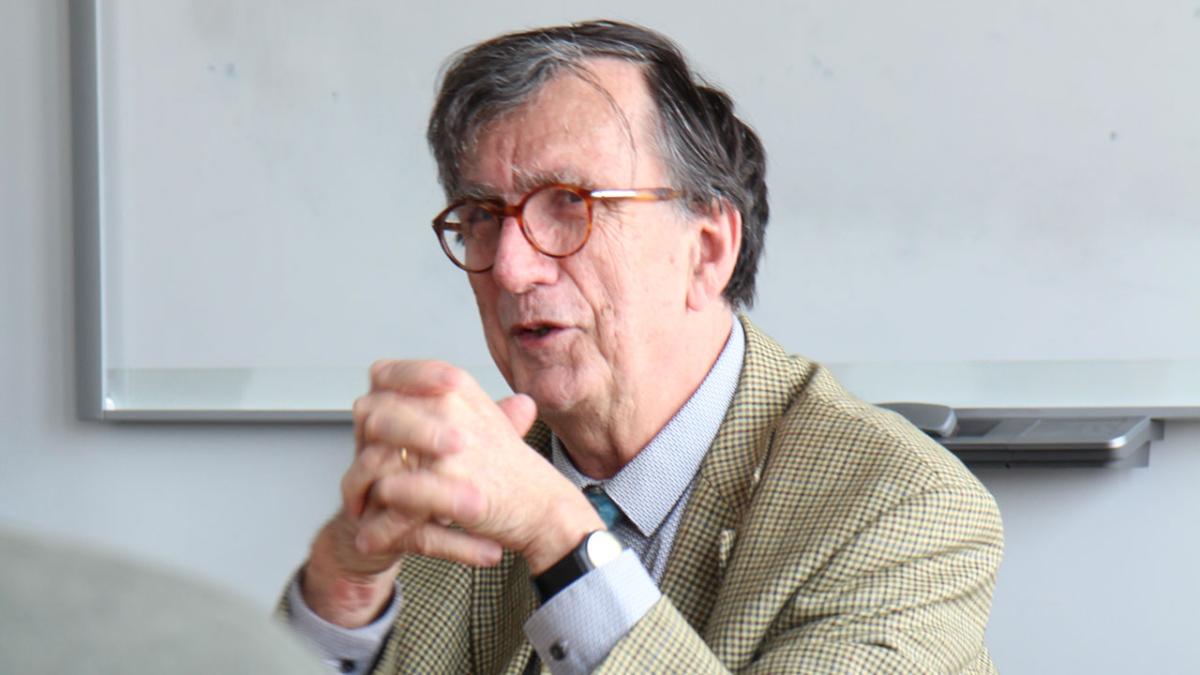 Archives numériques de Bruno Latour, photographie personnelle Portrait de Bruno Latour, pris de trois quart en train de parler, les mains jointes devant lui, devant un tableau d'une salle de cours.