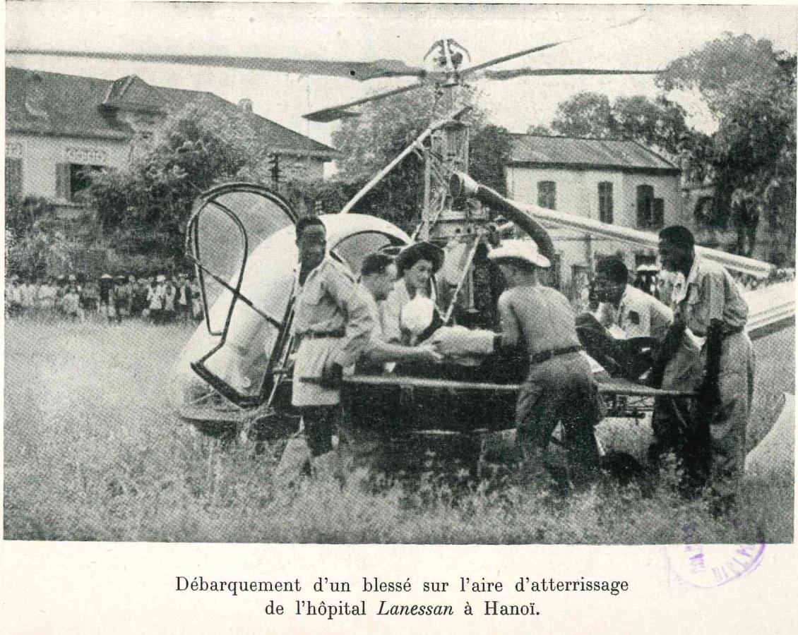 Photographie en noir et plan de Valérie André débarquant avec d'autres hommes un blessé sur une aire d'atterrissage avec en fond une hélicoptère 
