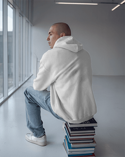 Modern Minimalist Hoodie Mockup Male Model In Blue Retro Style Design Sitting On Books Bright Glass Window Background