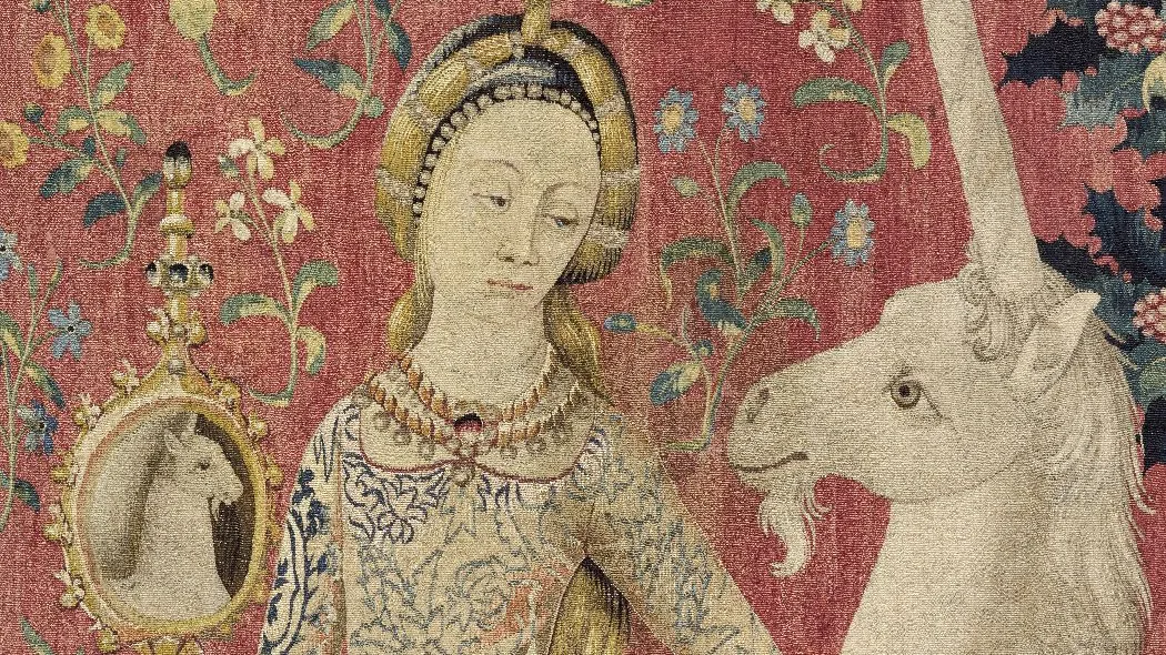 Tapisserie : une licorne se regardant dans un miroir tenu par une femme, sur fond rouge parsemé de fleurs. Vue de face.