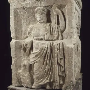 Bloque de piedra tallado: figura de cuerpo entero de un hombre de frente, vestido con una larga toga romana, sosteniendo una lanza. Fotografía frontal.