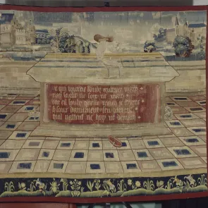 Tapisserie : main lançant une toupie sur une table, trois autres au sol, inscription à l’avant de la table. Vue de face.