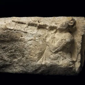 Bloque de piedra tallado: músico de perfil tocando una larga trompeta. Fotografía frontal.
