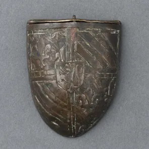 Boîte métallique en forme d’écu, ornée de fleurs de lis et de bandes diagonales, avec un blason central représentant un lion debout. Vue de face.