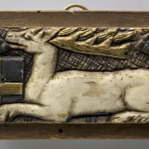 Plaque de bois sculptée et polychromée : un cerf couché de profil tenant entre les pattes un écu armorié. Vue de face.