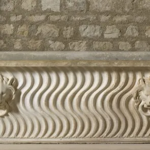 Cuve en pierre en forme de baignoire, décorée de courbes verticales et de deux têtes de lions tenant un anneau. Vue de face.