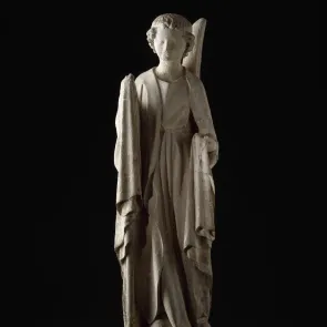Escultura en piedra de un ángel de pie con rostro redondo y vestido con ropa drapada. Vista frontal sobre fondo neutro.