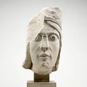 Escultura en piedra: cabeza femenina coronada, falta la parte superior derecha. Vista frontal.