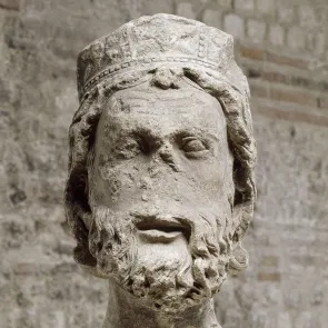 Cabeza esculpida y fragmentaria que representa a un hombre con barba rizada llevando un sombrero. Escultura en piedra de frente.