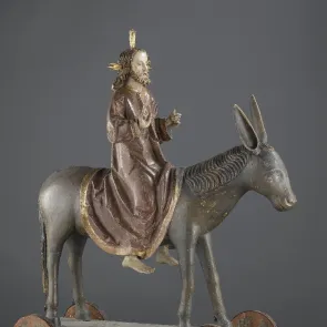 Escultura de madera pintada sobre ruedas que representa a Cristo montando un asno. Vista de perfil.