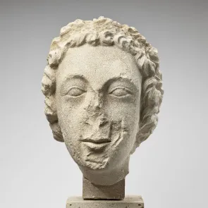 Tête sculptée en pierre vue de face représentant un personne aux cheveux bouclés retenus par un bandeau. Fond neutre. 