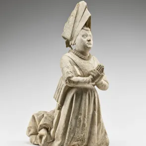 Escultura en piedra: mujer arrodillada en oración, con un vestido largo plisado y arrastre, y una alta cofia triangular. Vista de tres cuartos.