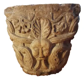 Capitel de piedra decorado con roleos de tres hebras que atraviesan una cabeza antropomorfa. Vista frontal.