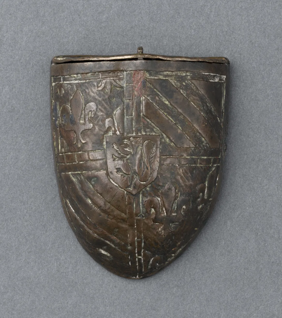 Boîte métallique en forme d’écu, ornée de fleurs de lis et de bandes diagonales, avec un blason central représentant un lion debout. Vue de face.