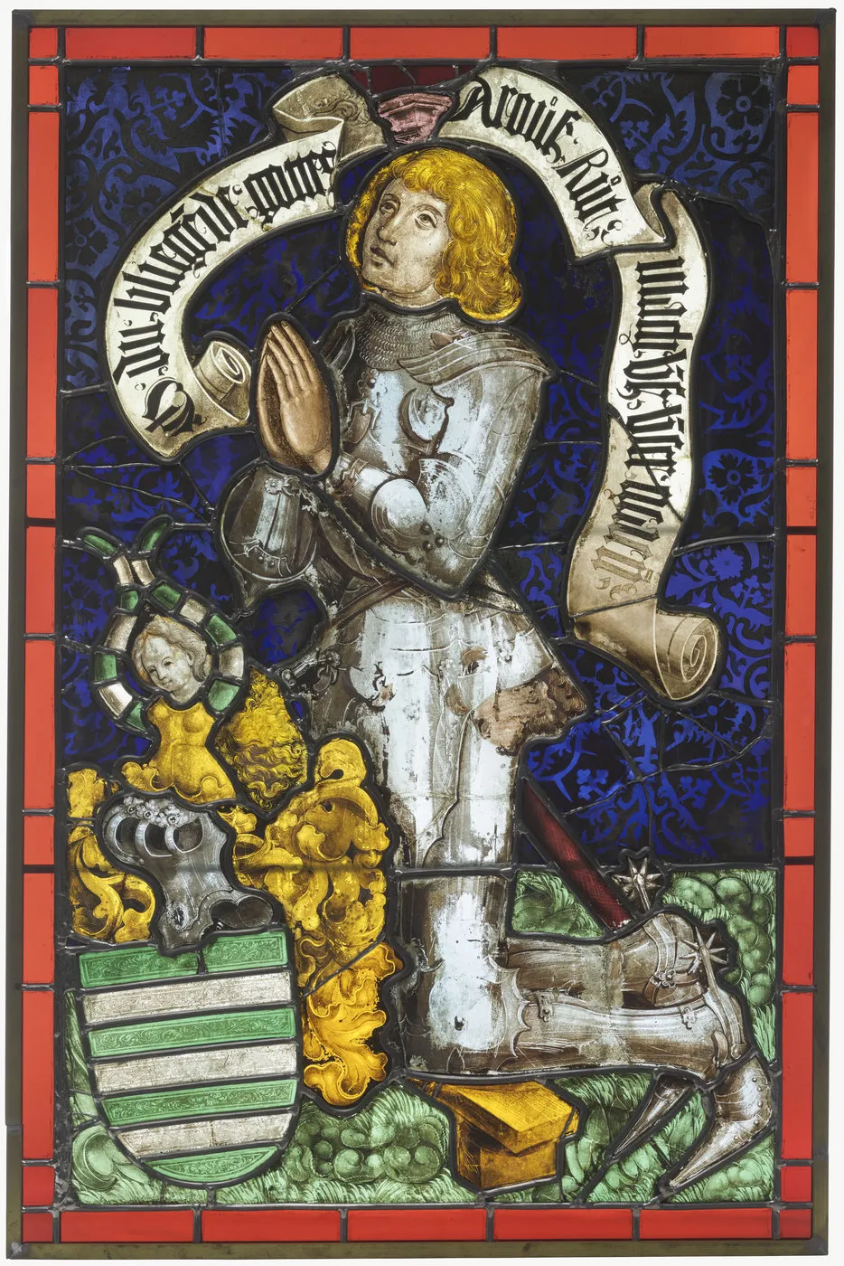 Vitral que representa a un hombre armado arrodillado en oración, con el rostro alzado. Vista frontal.