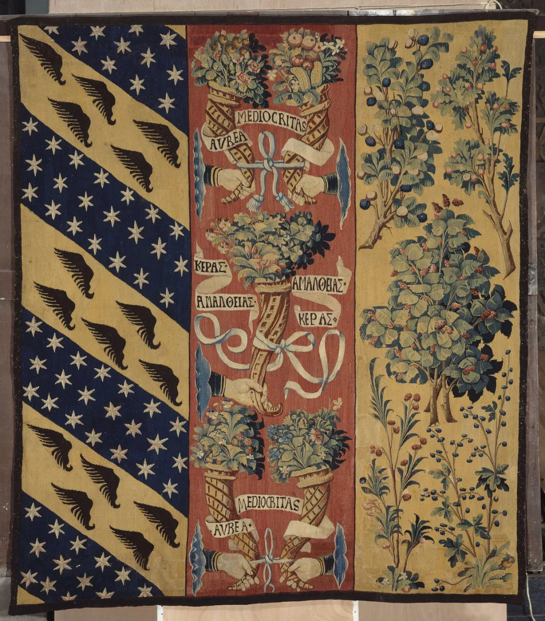 Tapisserie : mains tenant des cornes d’abondance au centre, motifs végétaux à droite, armoiries à gauche. Vue de face.