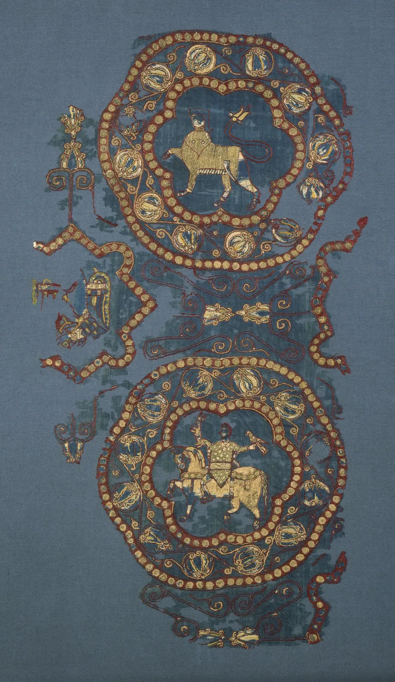 Fragment de tissu bleu brodé de rouge et or : médaillons avec un animal à tête humaine et un cavalier tenant un faucon et un lapin. Vue de face.
