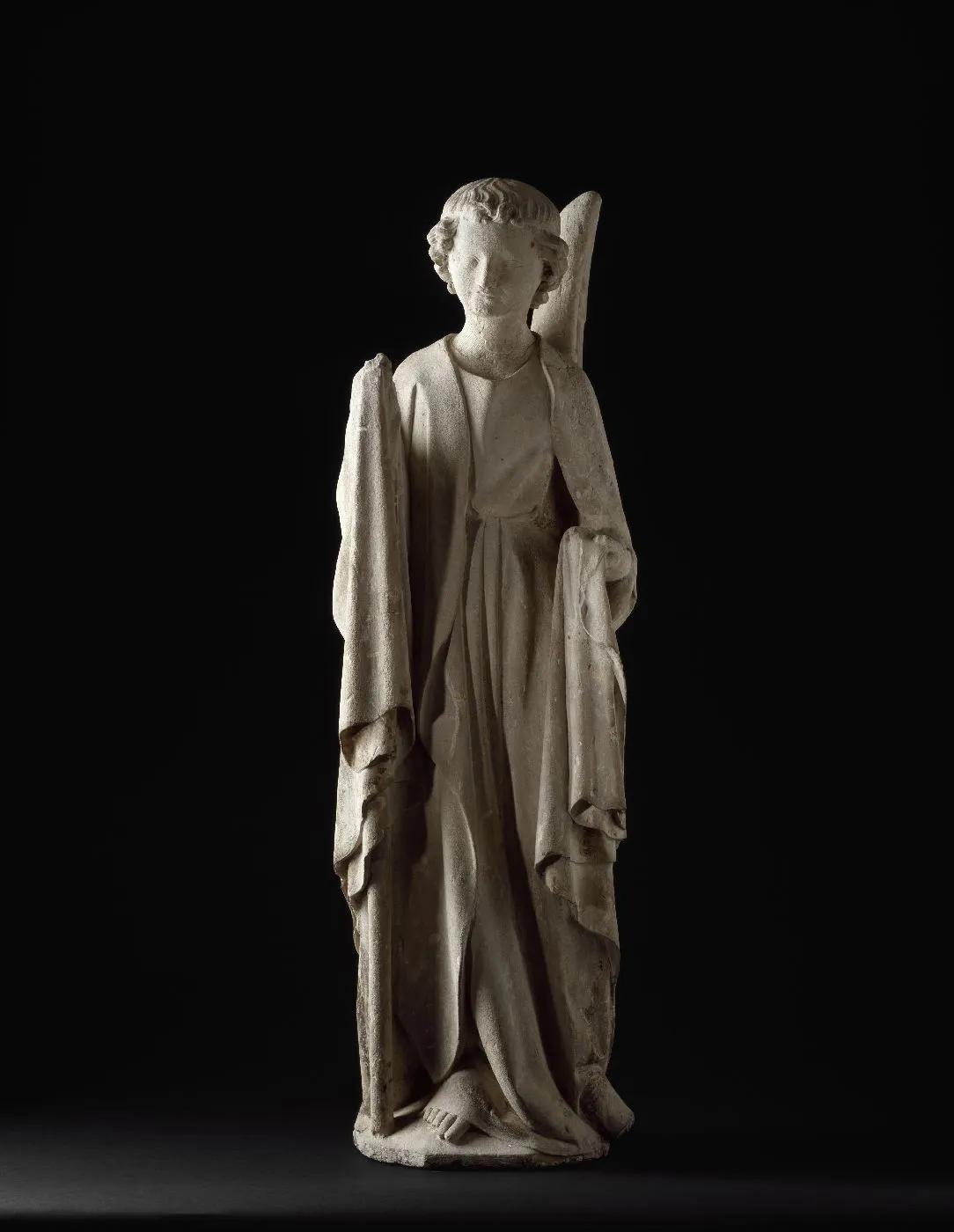 Sculpture en pierre d'un ange en pied au visage rond et vêtu d'un drapé. Vue de face sur fond neutre. 