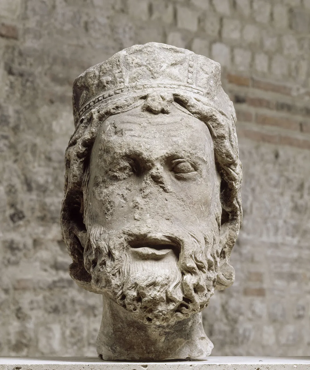 Tête sculptée et lacunaire représentant un homme à la barbe bouclée portant un chapeau. Sculpture en pierre de face. 