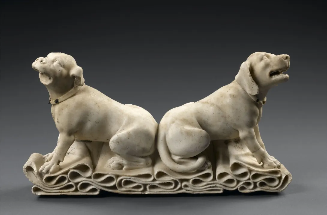 Sculpture en pierre : deux chiens assis dos à dos sur un tissu plissé, regardant vers le ciel. Photographie de profil.