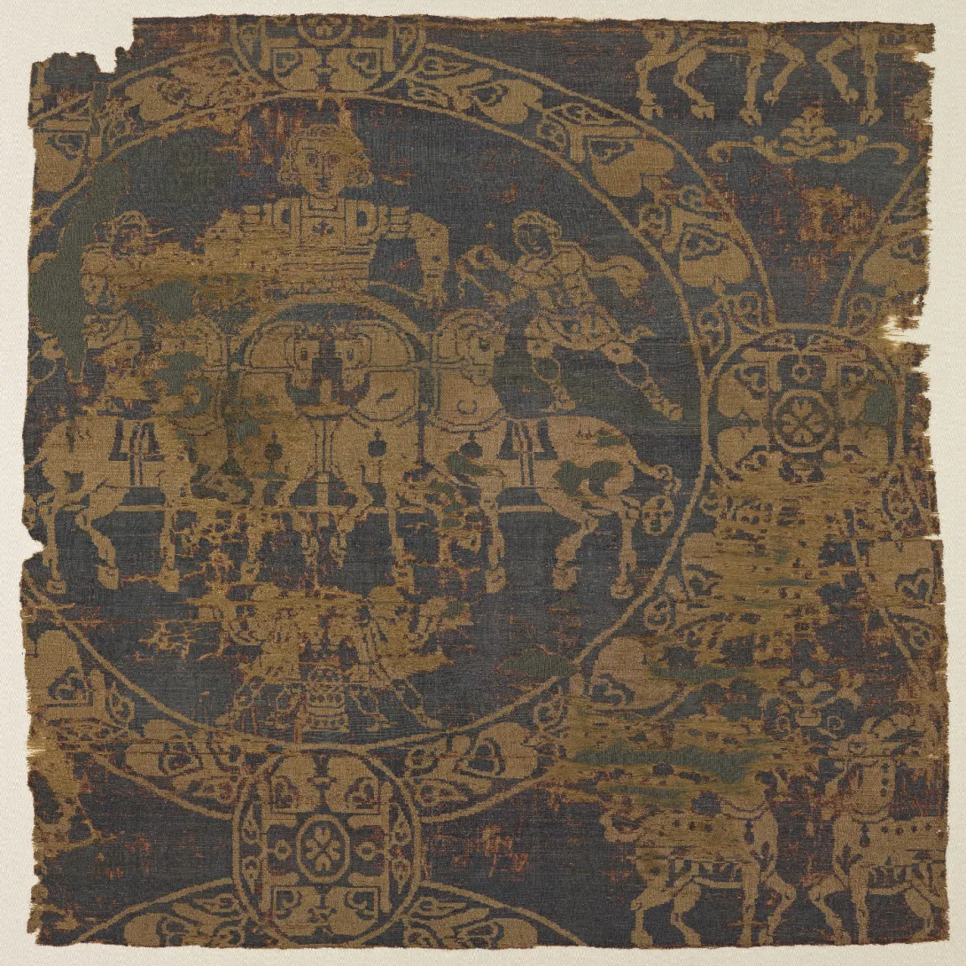 Fragment de tissu marron. Dans un médaillon se trouve une scène avec un personnage sur un char porté par des chevaux. 