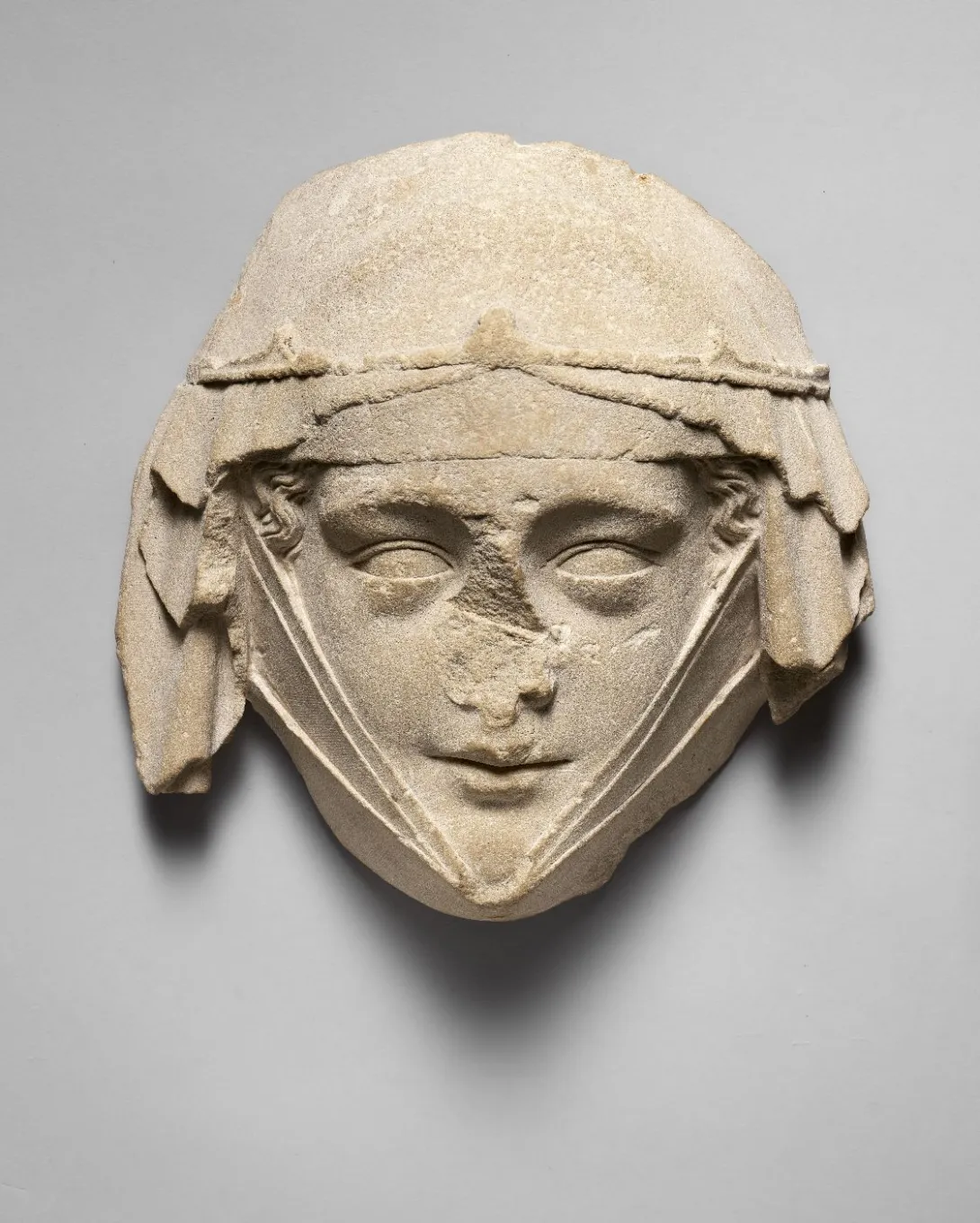 Sculpture en pierre représentant un visage féminin (nez manquant), portant un voile retenu par une fine couronne et une guimpe. Vue de face.