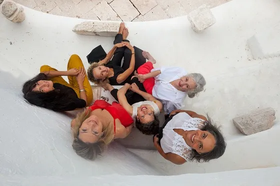 Six femmes sont vues de haut, le visage tourné vers l'appareil photo. Quatre sont assises et deux debout dans un décor minéral.