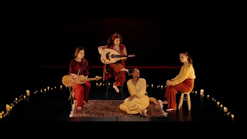 Quatre femmes sont sur scène, sur fond noir. Deux jouent d'un instrument. Elles sont assises dans un demi-cercle de bougies. 