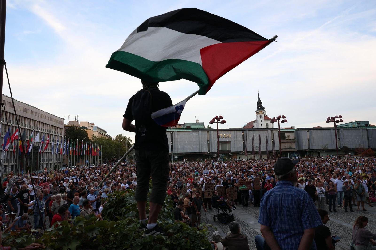 Človek vihra s palestinsko zastavo, v ozadju protest na Trgu republike.