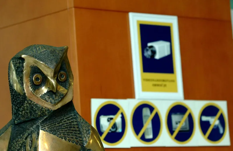 Kip Sove, v ozadju nalepke za različne prepovedi in opozorilo, da se prostor snema.