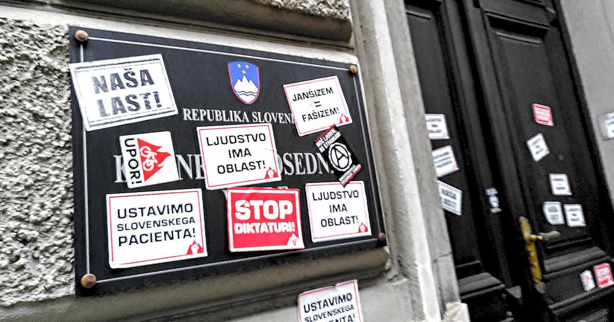Črnobela fotografija table Kabinet predsednika vlade RS, ki je polepljena z rdečebelimi protestniškimi nalepkami (naša last, ljudstvo ima oblast, stop diktaturi, ustavimo slovenskega pacienta, janšizem = fašizem).