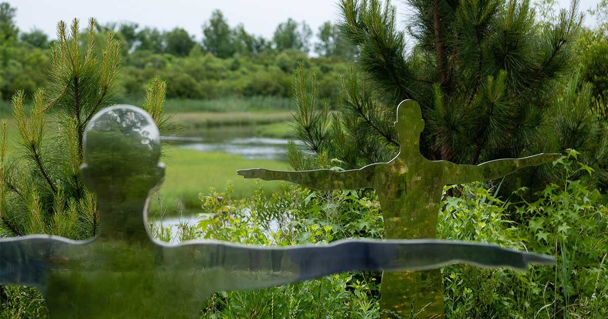 Zelena pokrajina z vodo, močvirjem in bujnim zelenim rastjem v ozadju, v ospredju sta dve skulpturi človeške oblike iz materiala, ki reflektira okolje. Obliki se zlijeta z zelenim okoljem.