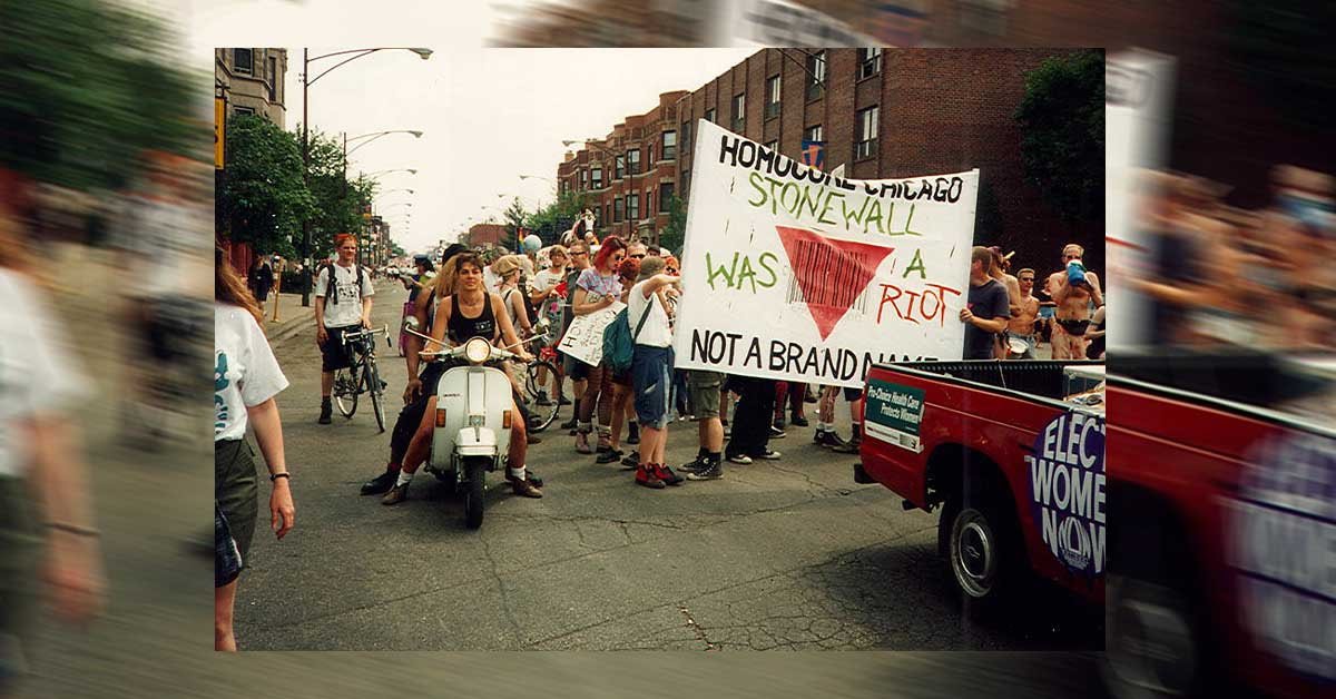 na fotografiji je prizor povorke, ki hodi za transparentom, na katerem piše Stonewall was a riot not a brand name