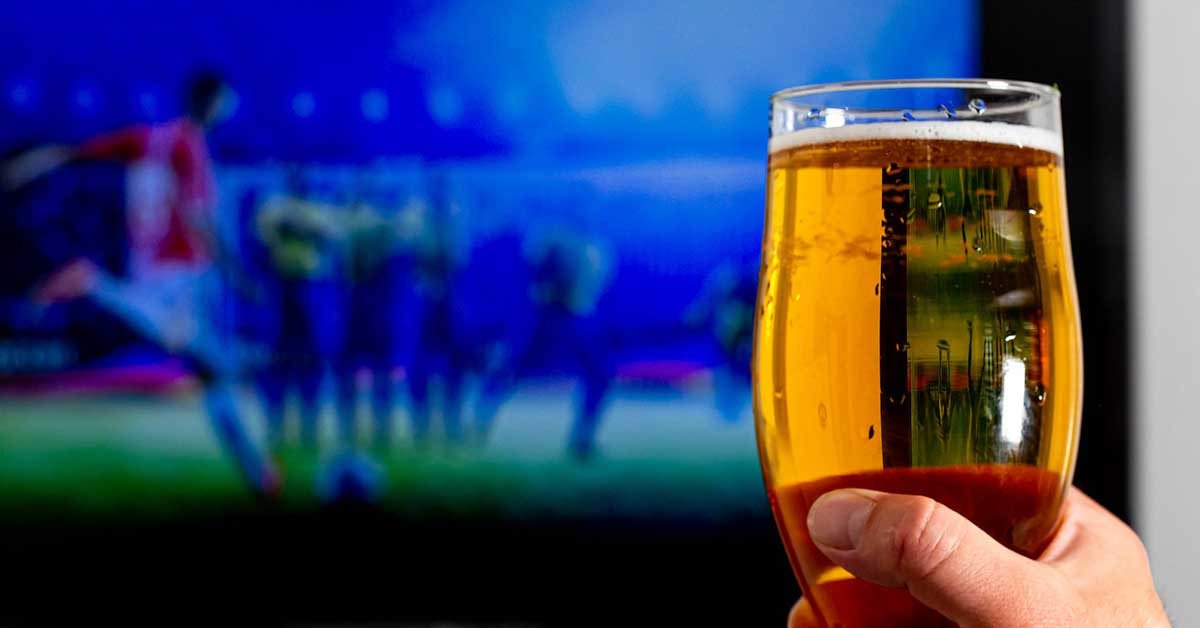 Roka, ki drži kozarec piva, v ozadju pa se predvaja nogometna tekma.