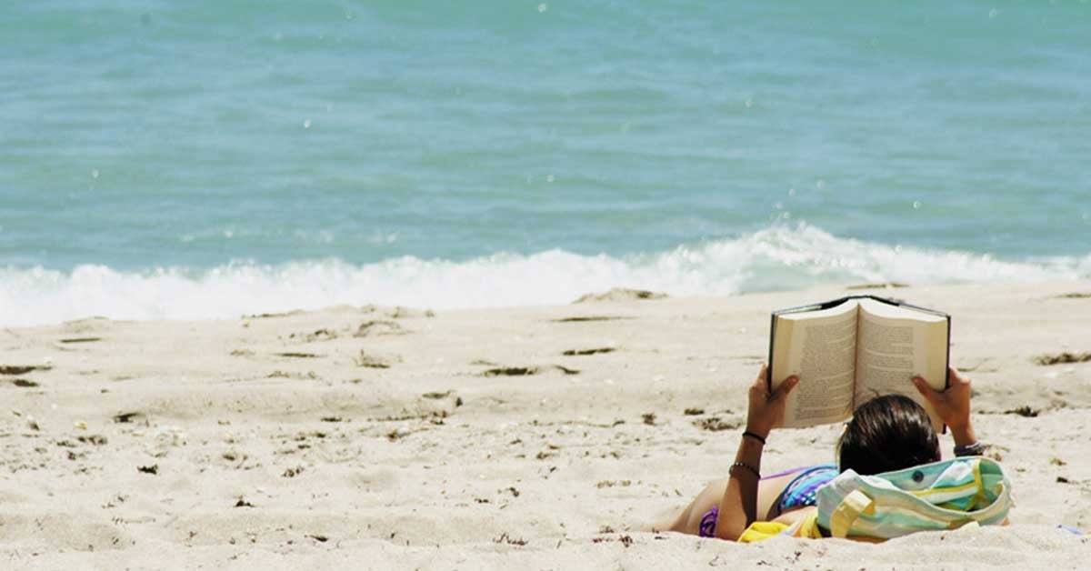 Oseba na peščeni plaži bere knjigo.