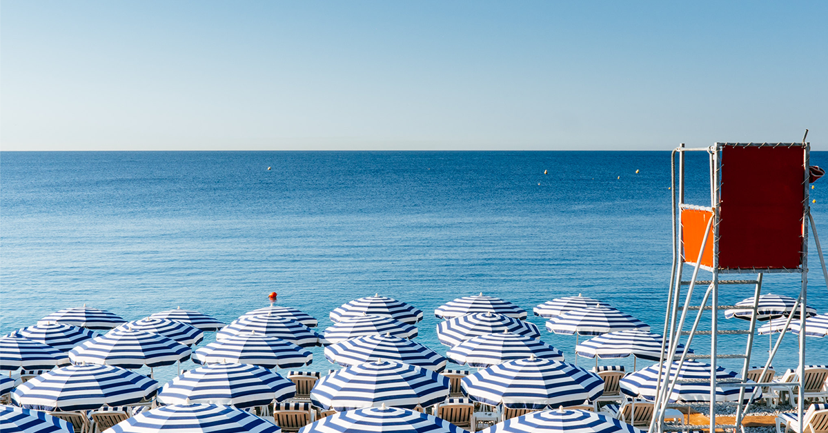 Fotografija modro-belih senčnikov in reševalnega stolpa na plaži s pogledom na morje.